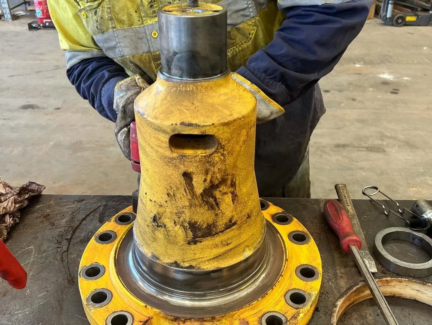 A worker in hi-vis and gloves steadies a tall yellow cast component standing on a bolted yellow flange on a steel workshop bench, with a chisel, a steel ring and a screwdriver beside it.