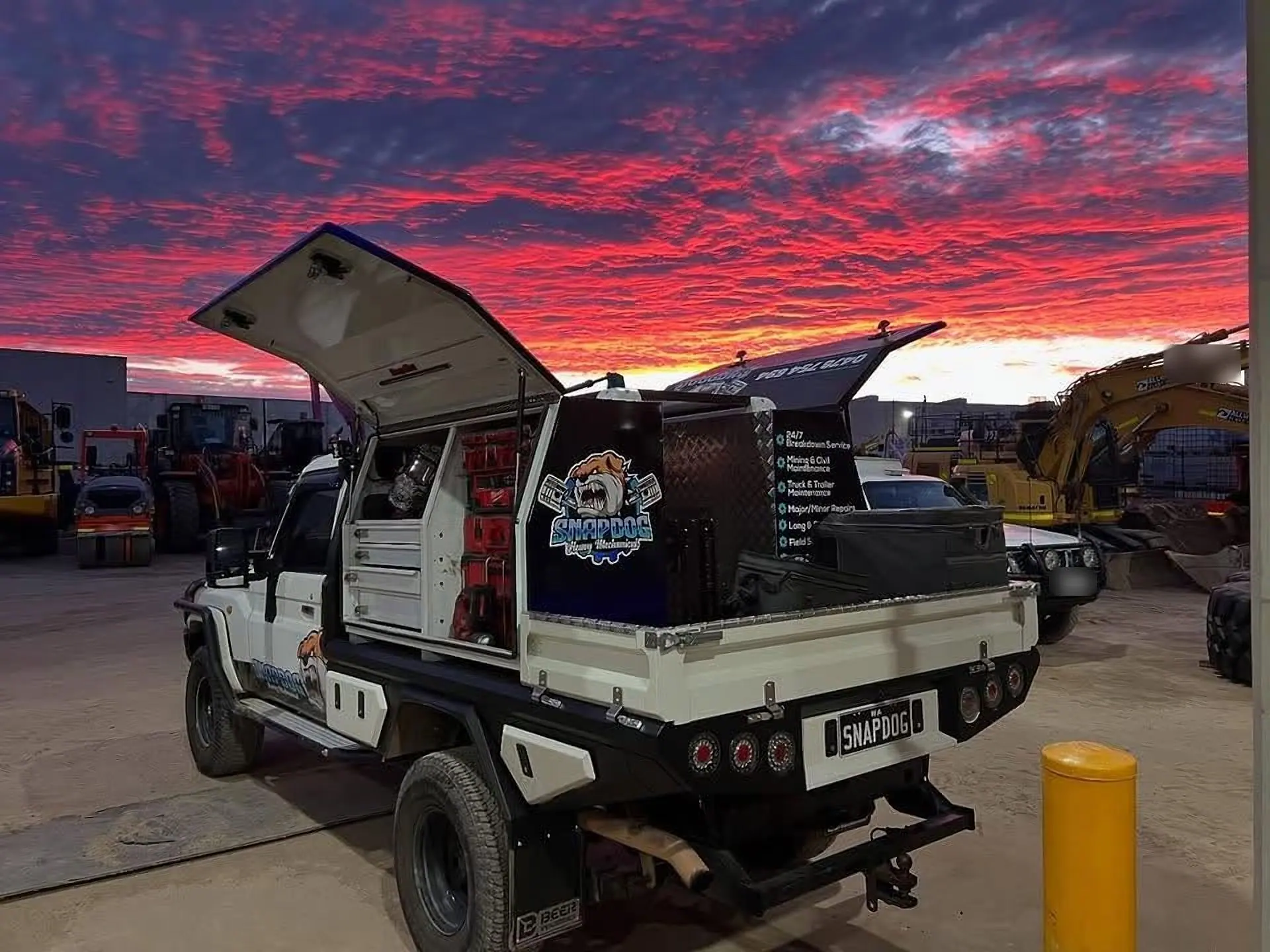SnapDog's tray-back with its canopy doors raised against a red dusk sky, with excavators and loaders parked behind.