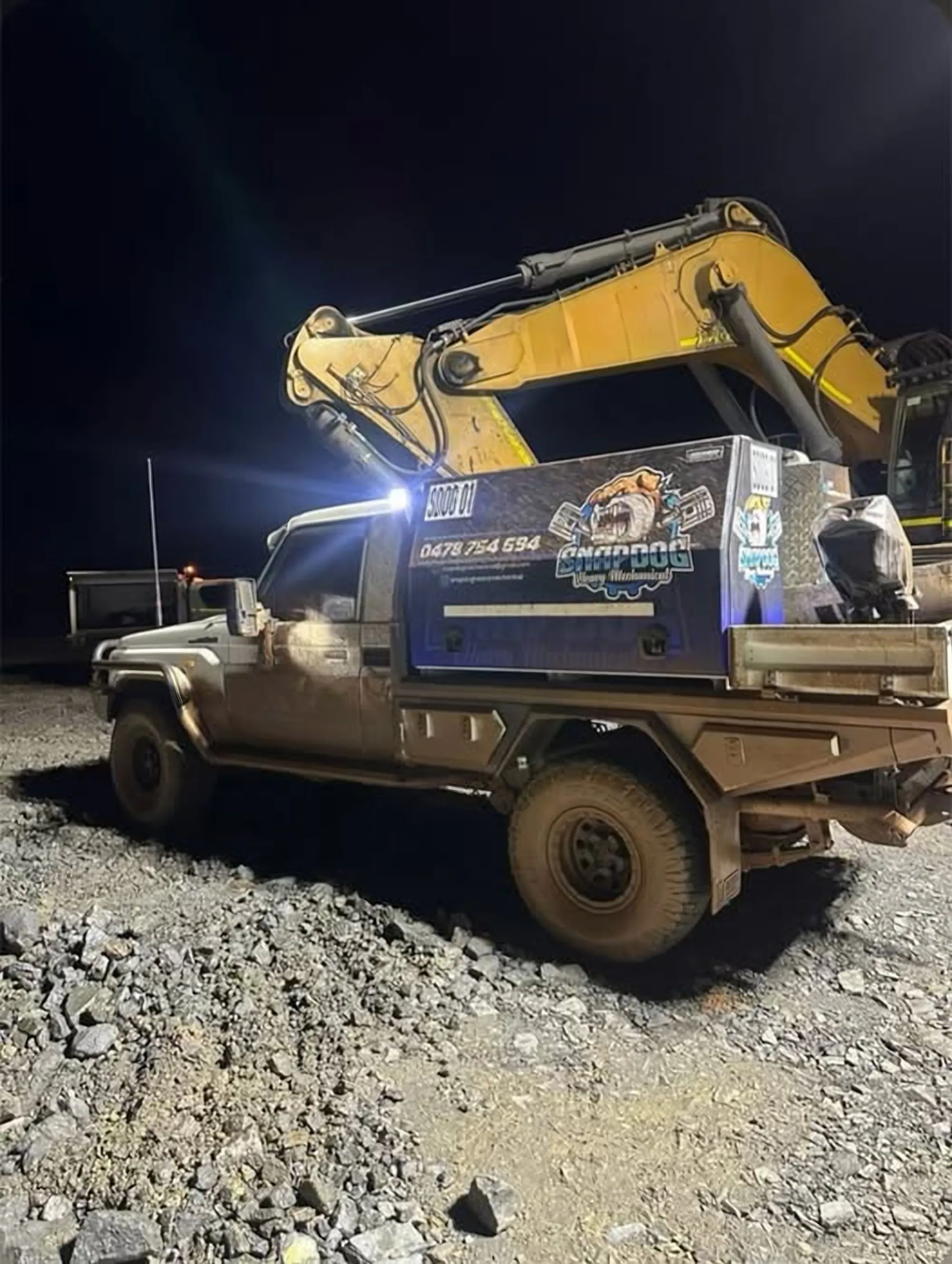 SnapDog's tray-back at night with work lights on and an excavator boom lit up behind it.