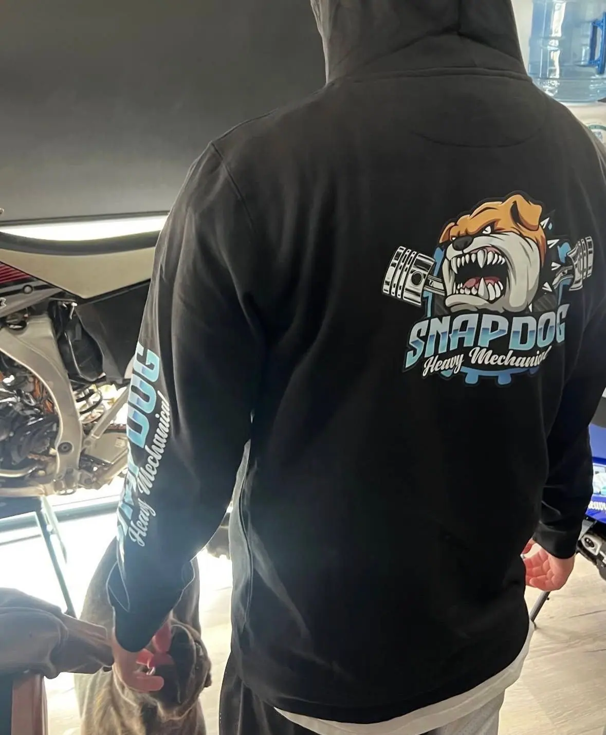 The back of a black SnapDog hoodie with the bulldog logo across the shoulders and the SnapDog Heavy Mechanical name down the sleeve.