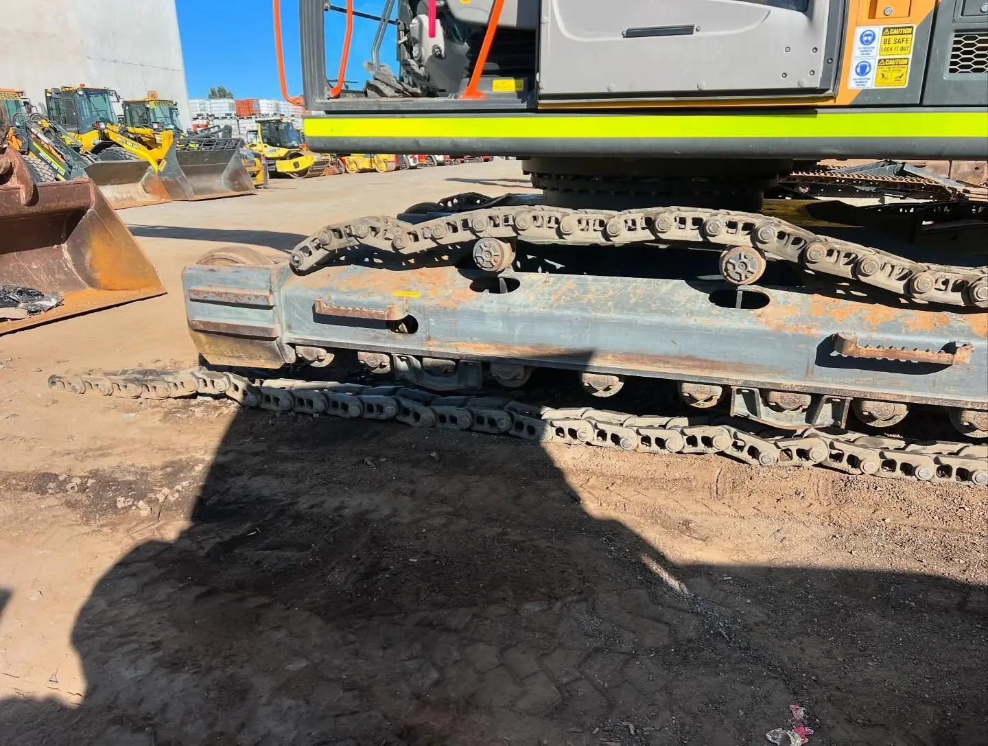 A steel track chain and yellow undercarriage laid out in the sun beside an excavator.