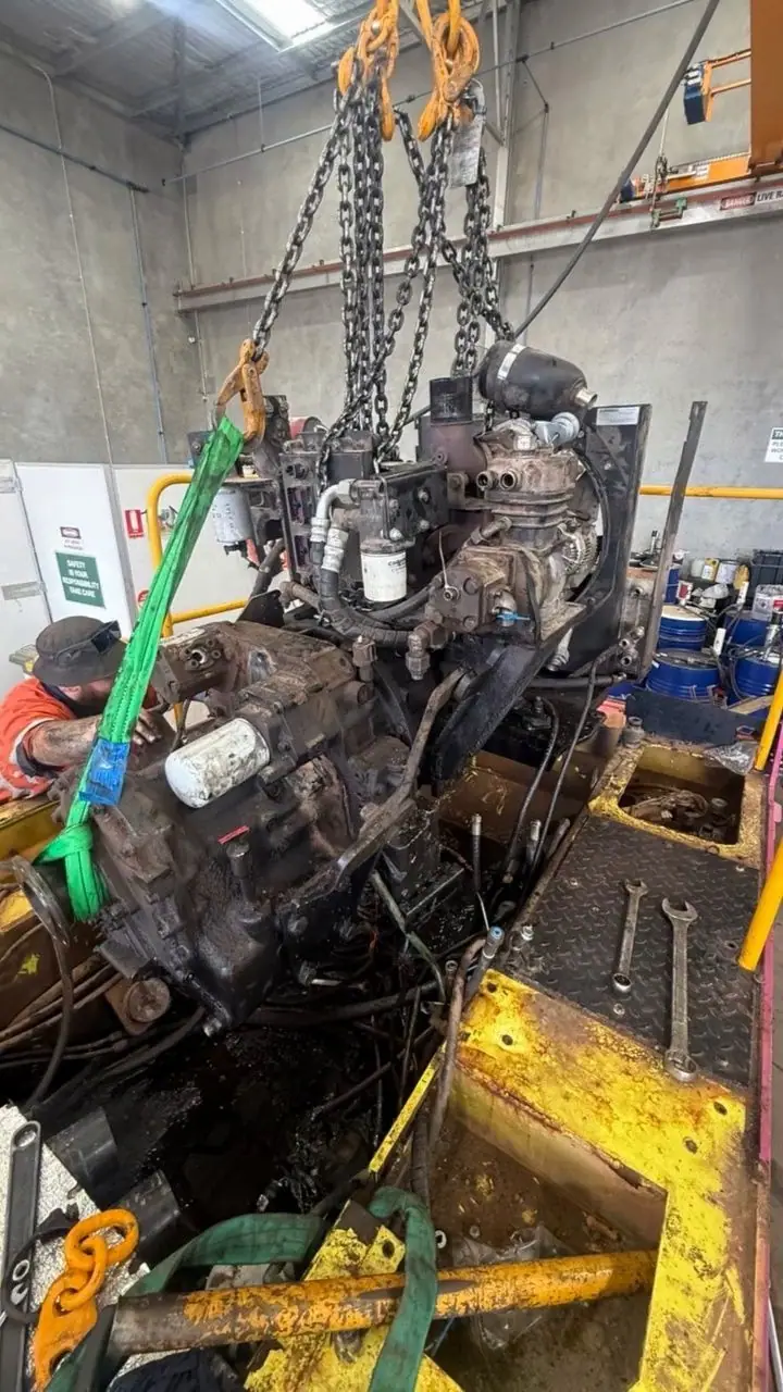 A diesel engine hanging on chains above a yellow machine chassis with spanners resting on the deck plate.