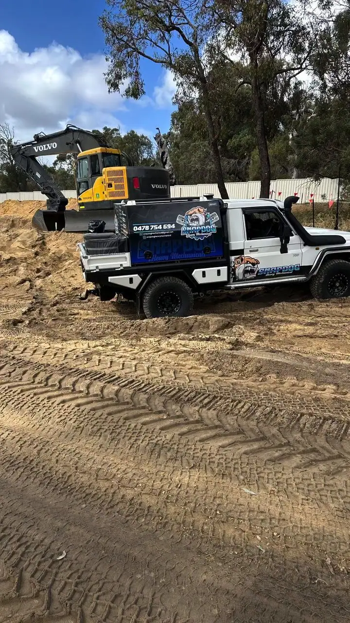 SnapDog's white Land Cruiser tray-back on a sandy civil site in front of a yellow Volvo excavator, with tyre tracks in the foreground.